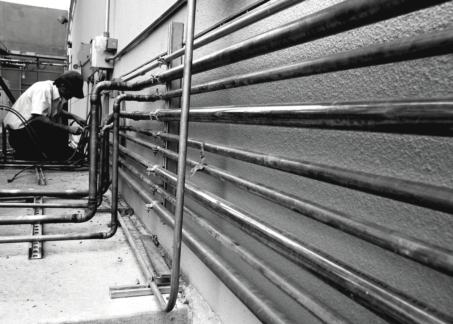 Man working on metal pipes attached to a wall outdoors. Black and white.