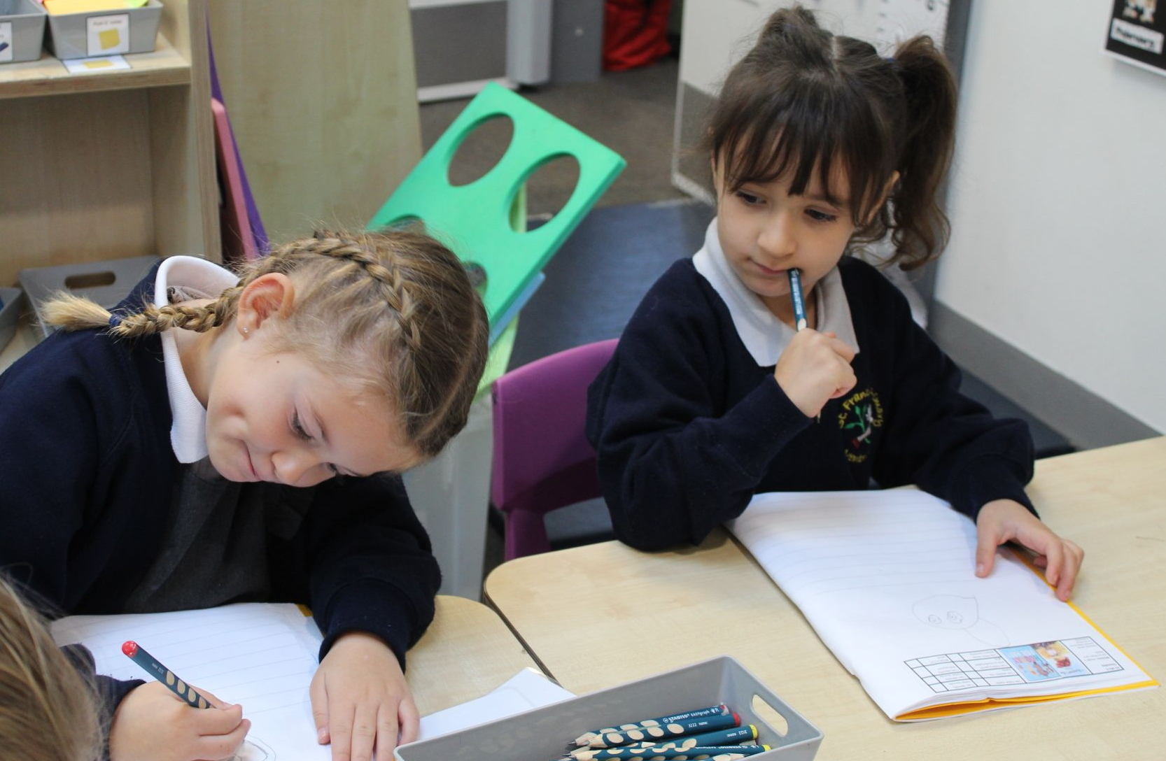Two young girls in uniform writing at desks; one thoughtful, the other focused, in a classroom setting.