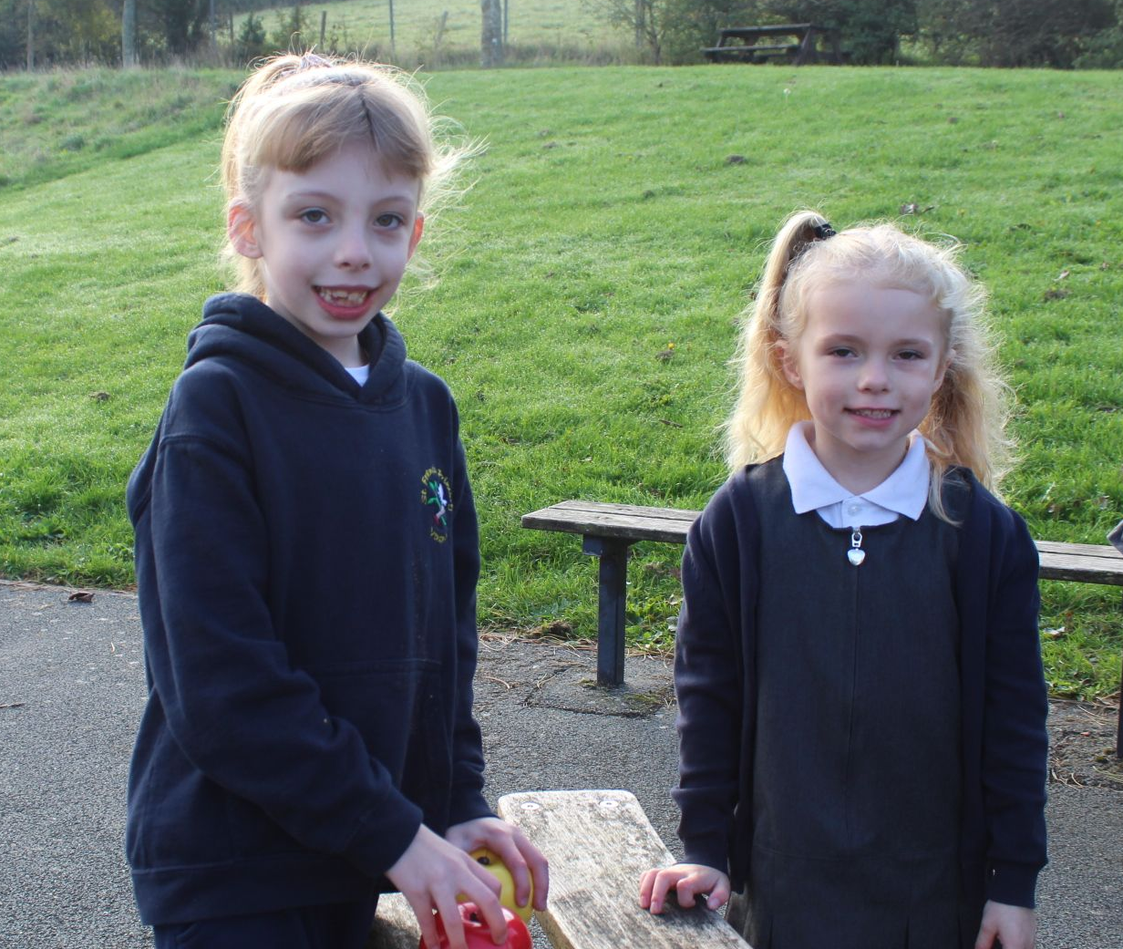 Two young girls in school uniforms stand outdoors, one smiling with a ponytail, the other with mouth open.
