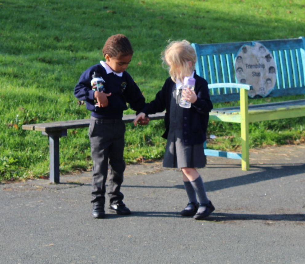 Two young students in uniform holding hands, walking in front of a park bench.