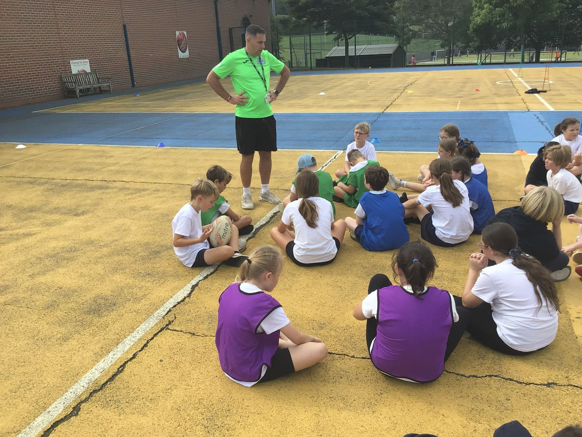 A group of children in school uniforms sitting in a circle, listening to an instructor on a sports field.