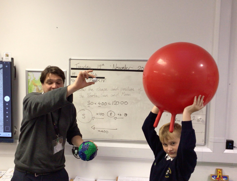 A teacher and a boy are holding a spacehopper in a classroom to compare sizes between earth and sun