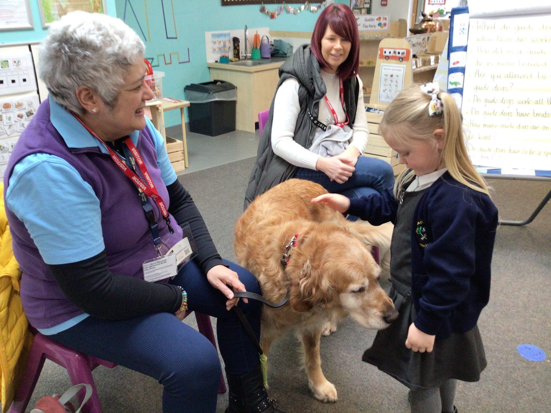 Woman and child pet a golden retriever in a classroom while an older woman sits nearby.