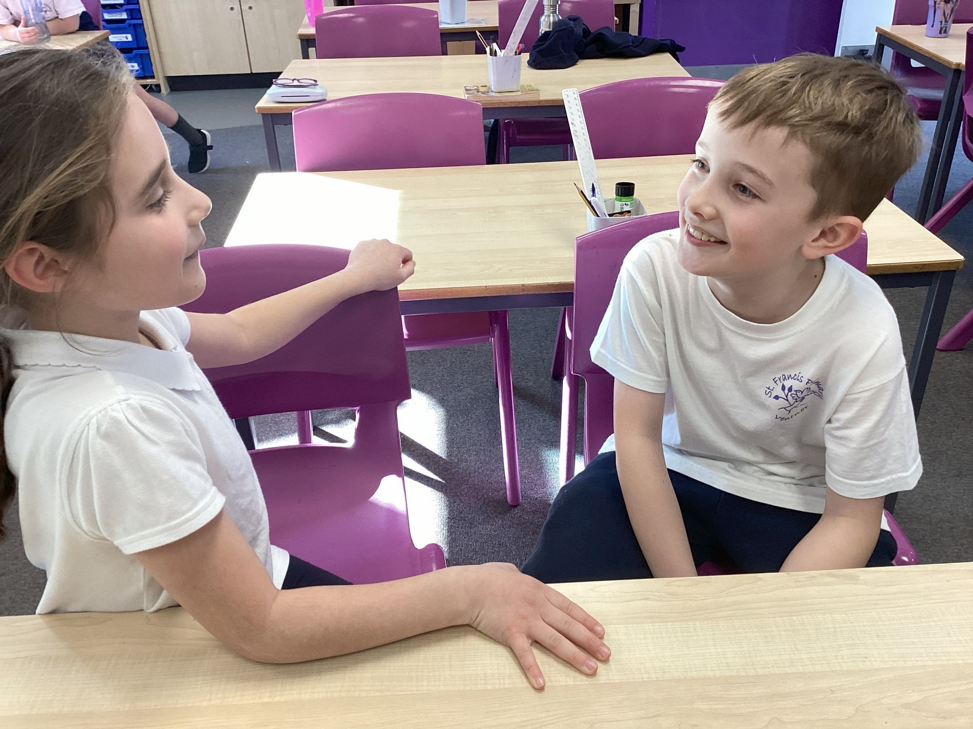 Two children sitting at a classroom table, smiling and talking across from each other.