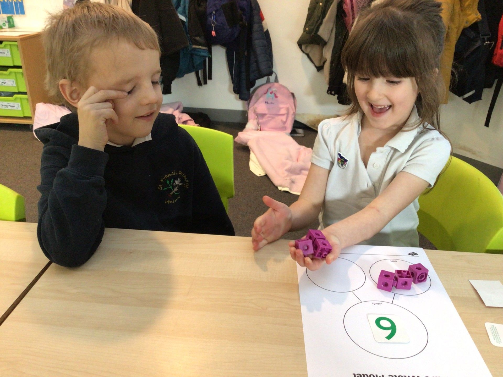 Two children at a table, counting purple cubes. One looks on, the other smiles and holds cubes over a worksheet.