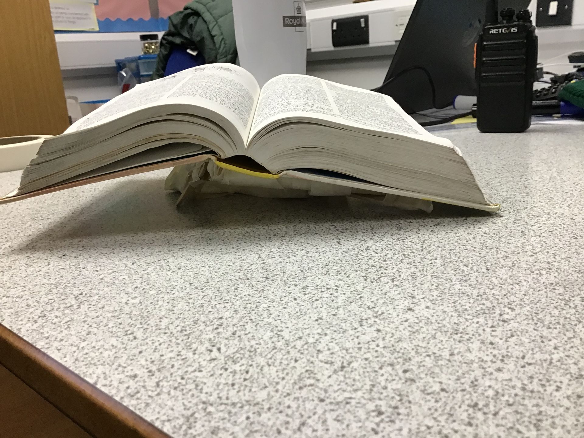 Four children in school uniforms, gathered around an open book on a desk. Two stand behind, two seated in front.