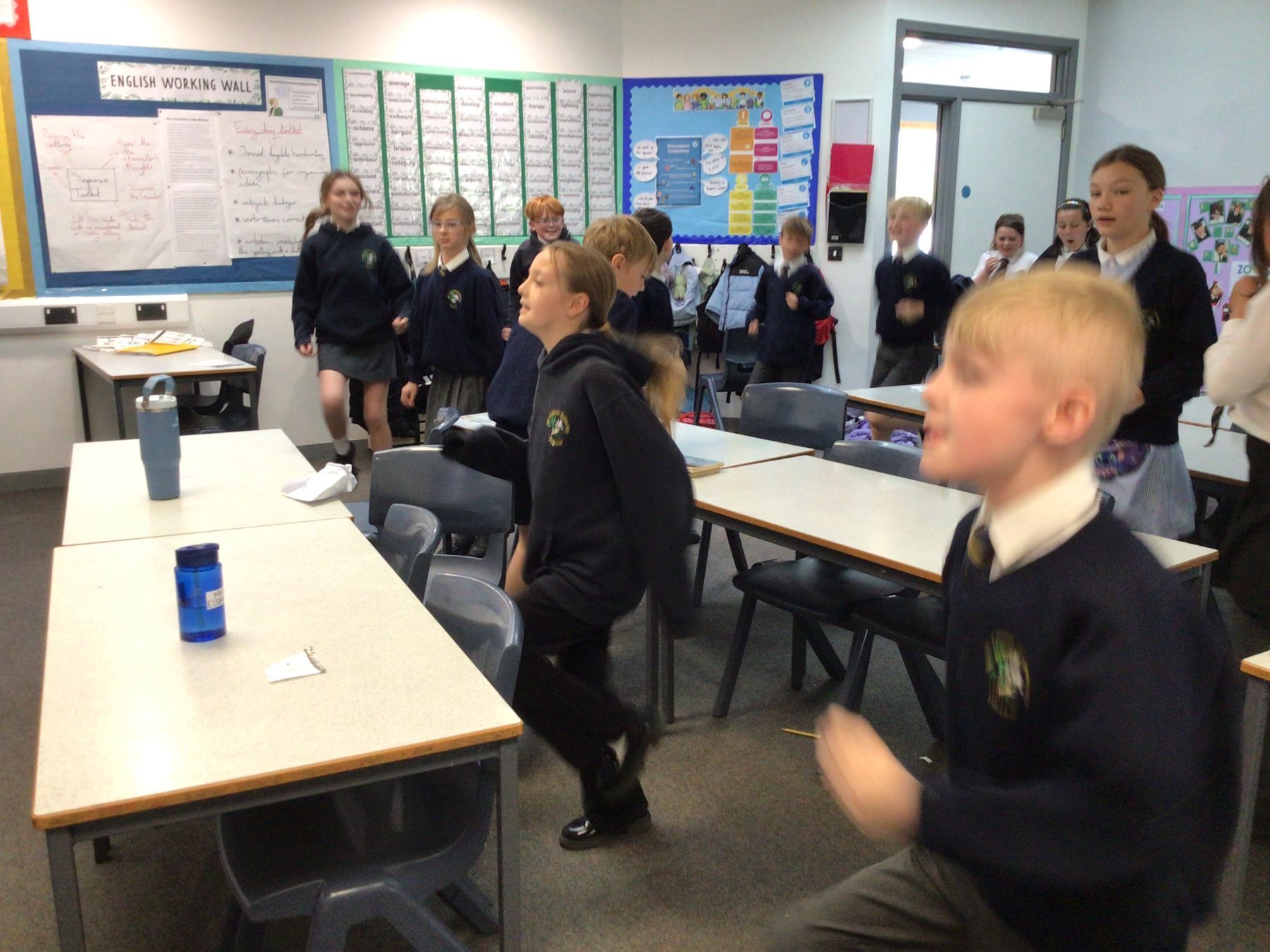 Students in school uniforms jumping in a classroom during an activity.