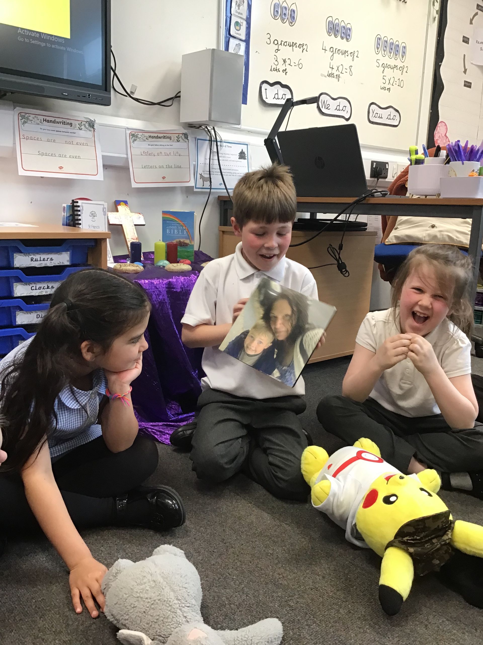 Children sitting on a classroom carpet, smiling and laughing with stuffed toys and a book.
