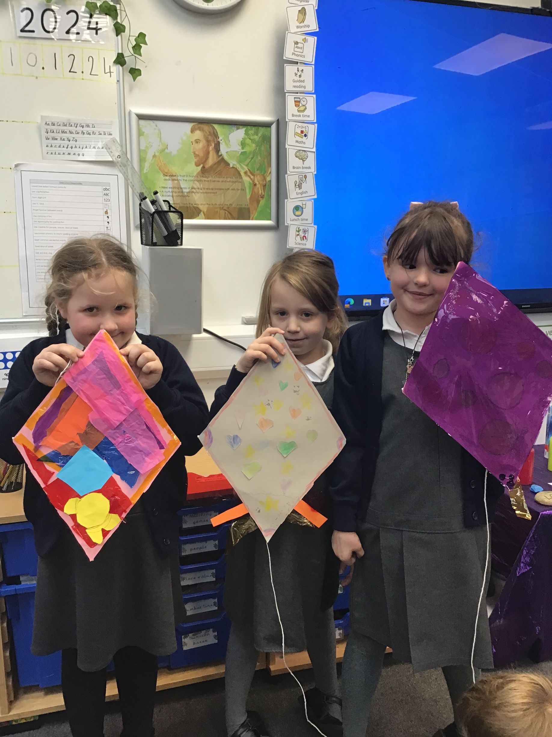 Three young girls are holding kites in a classroom.