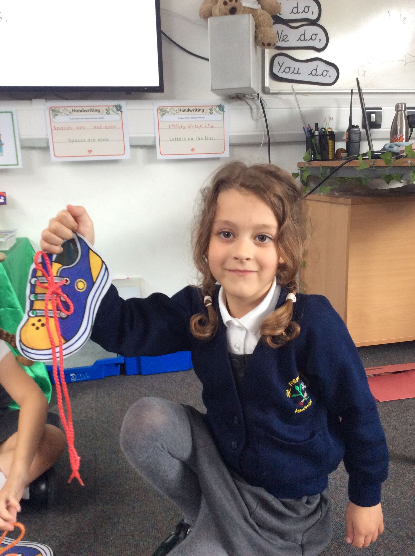 A little girl in a school uniform is holding a cardboard shoe cut-out with tied laces