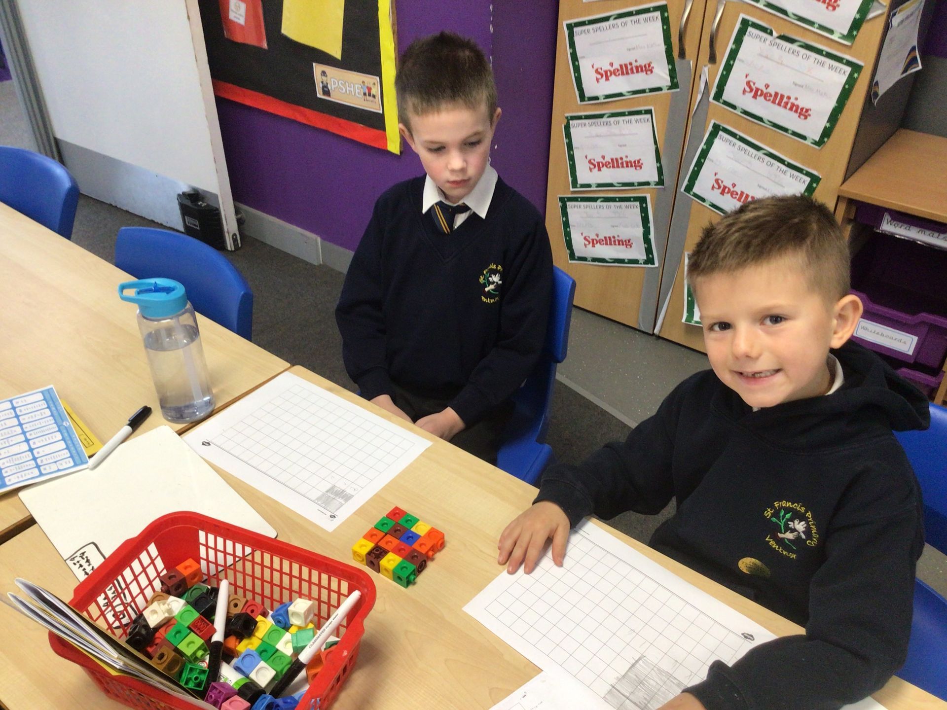 Two schoolboys sit at a desk with grid paper and building blocks. One smiles at the camera. Classroom setting.