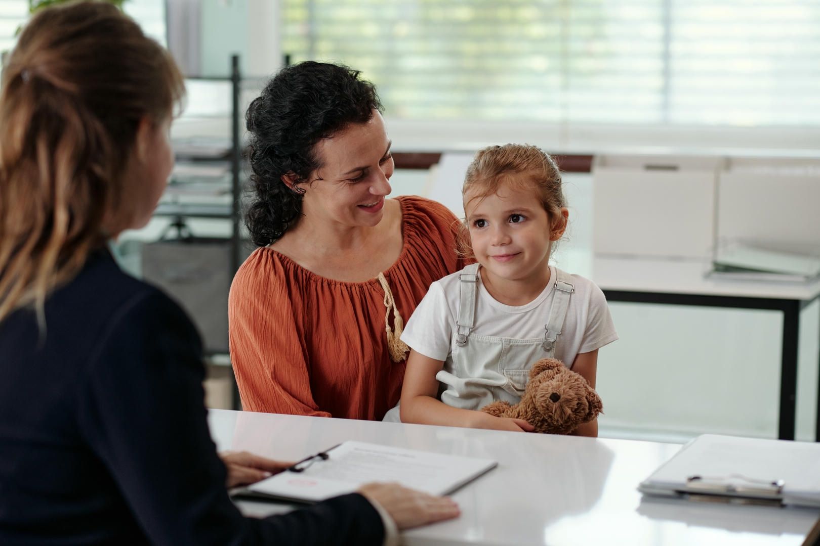 A woman and a little girl are sitting at a table with a teddy bear.