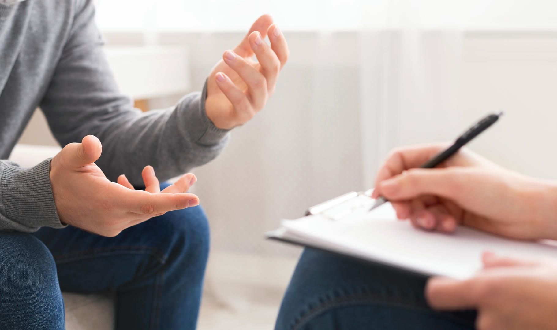 A man is sitting on a couch talking to a woman who is writing on a clipboard.