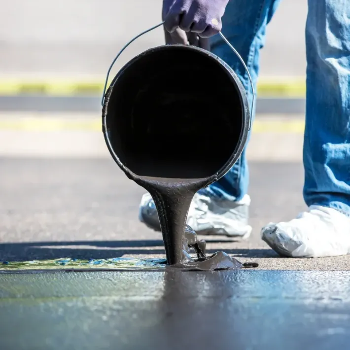 Person pours black asphalt from a bucket onto a road surface.