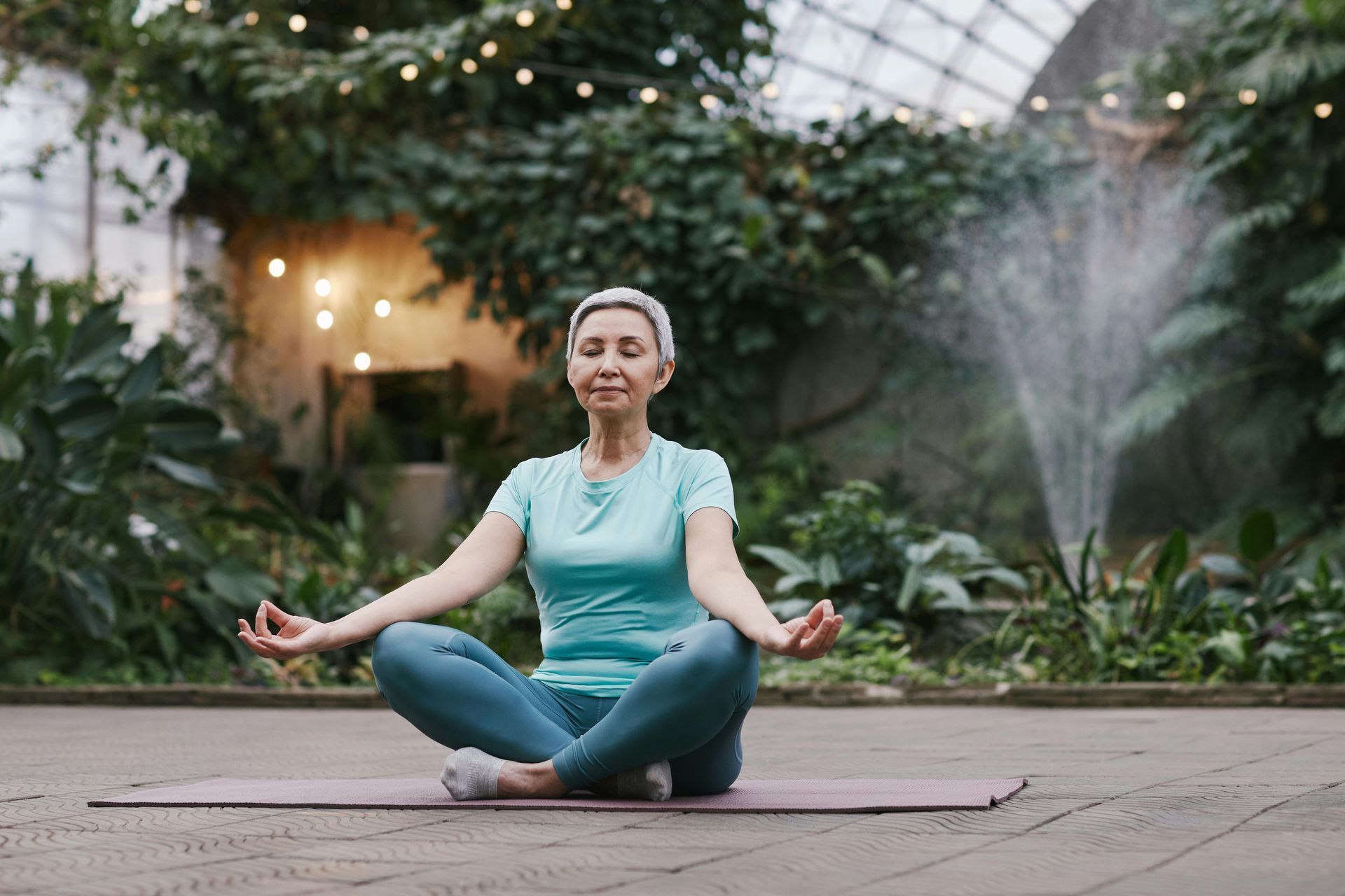 An elderly woman is sitting in a lotus position on a yoga mat.