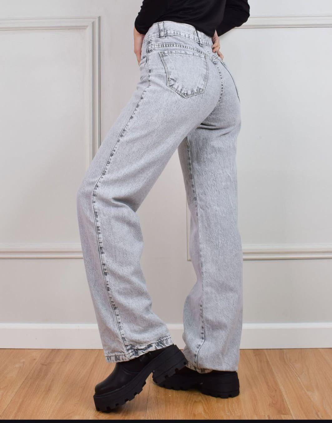 Mujer con jeans de mezclilla claros y zapatos de plataforma negros, de pie en un interior con paredes blancas.