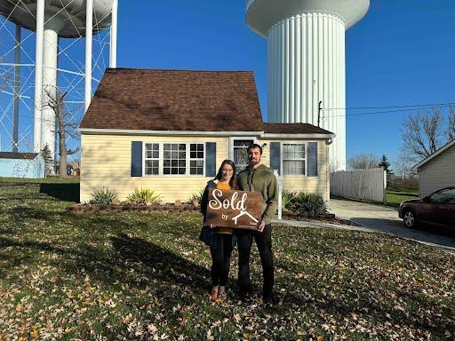 A man and a woman are standing in front of a house holding a sold sign.