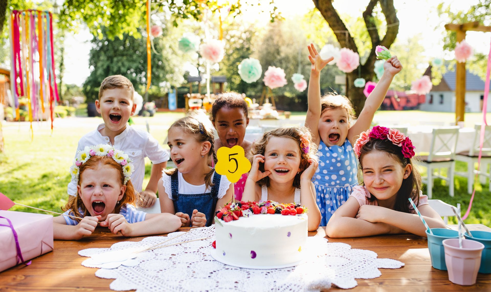 Children at birthday party, cheering, cake with strawberries, outdoor setting.