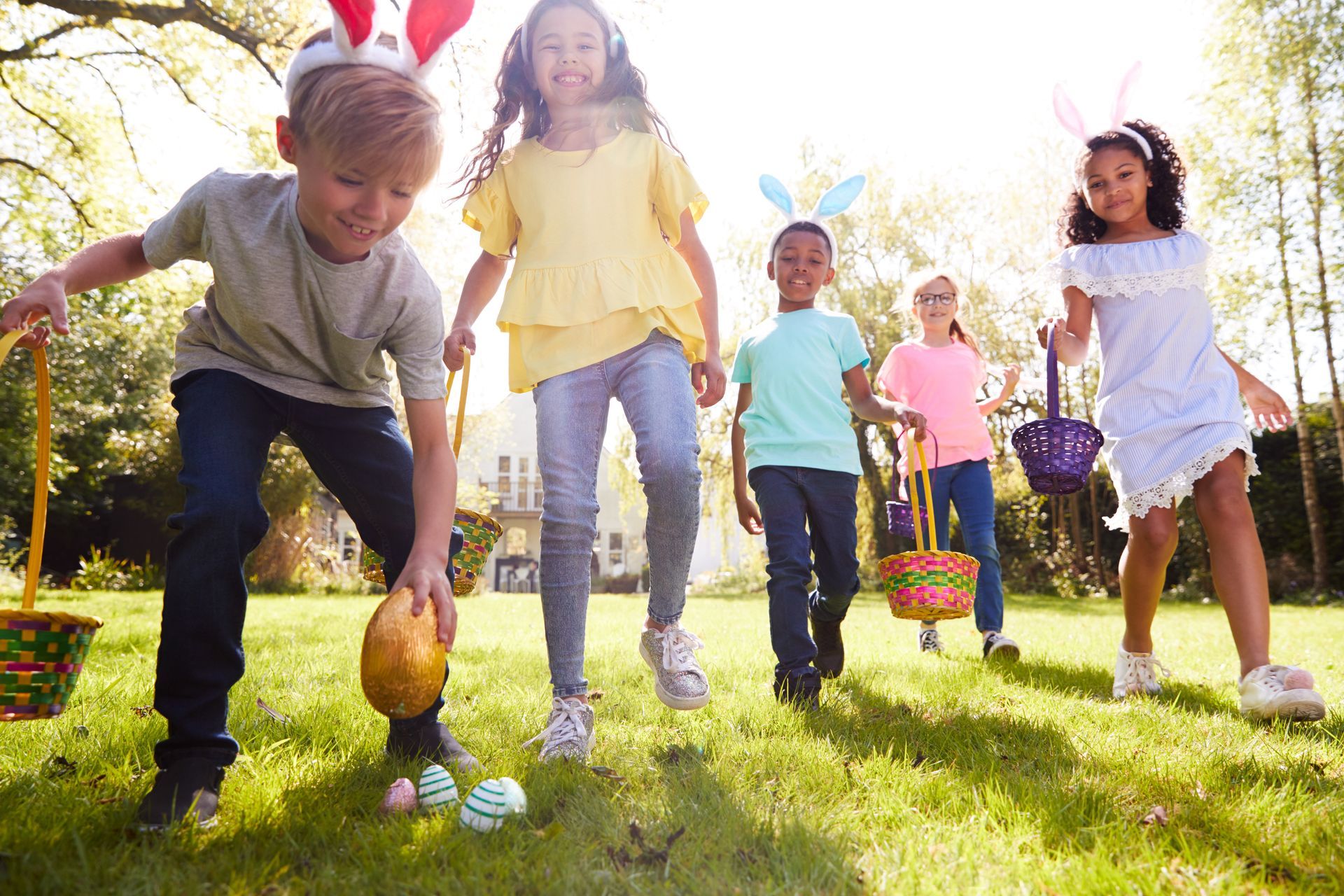 Children with bunny ears hunt for Easter eggs in a sunny yard, holding baskets.