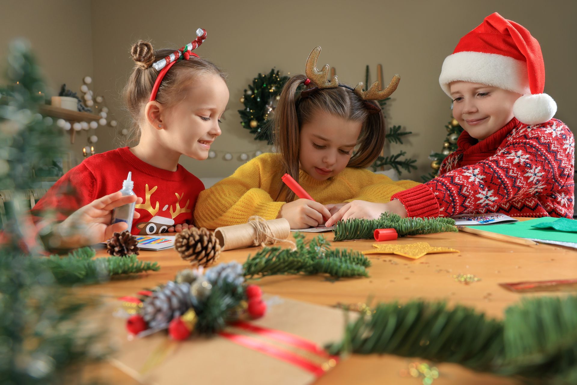 Children craft Christmas decorations at a table. One draws, others glue. Festive sweaters and headbands.
