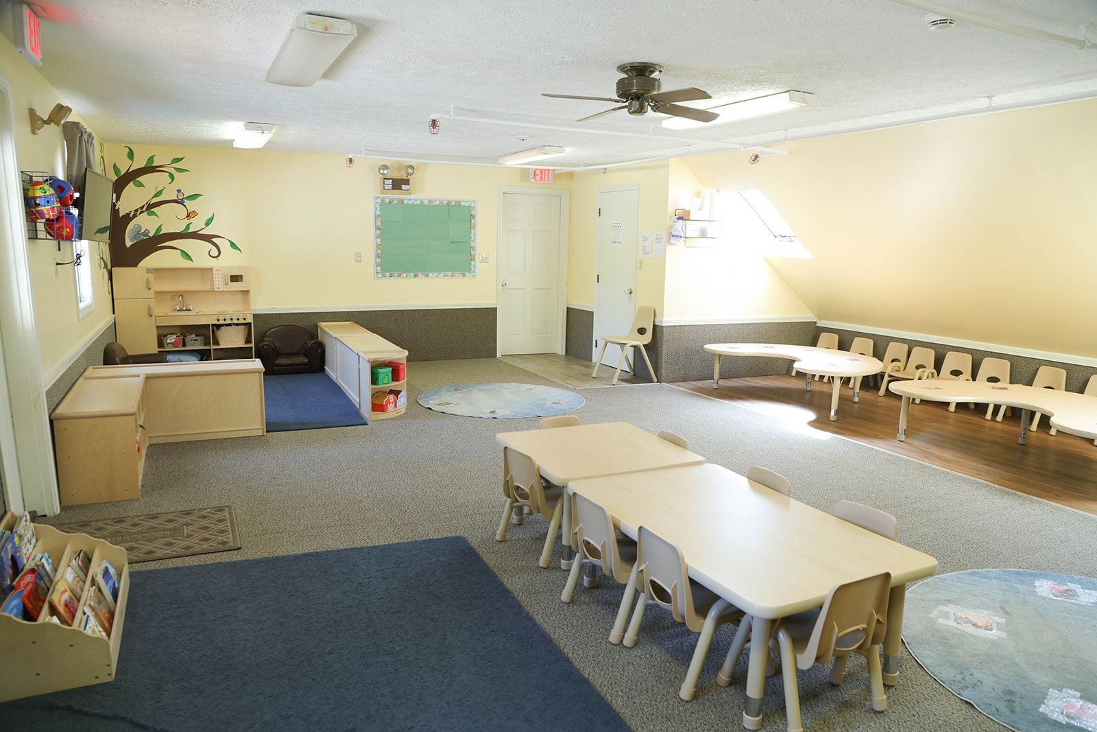 Daycare classroom, tan walls, tables, chairs, blue rug, shelves, tree mural, natural light.