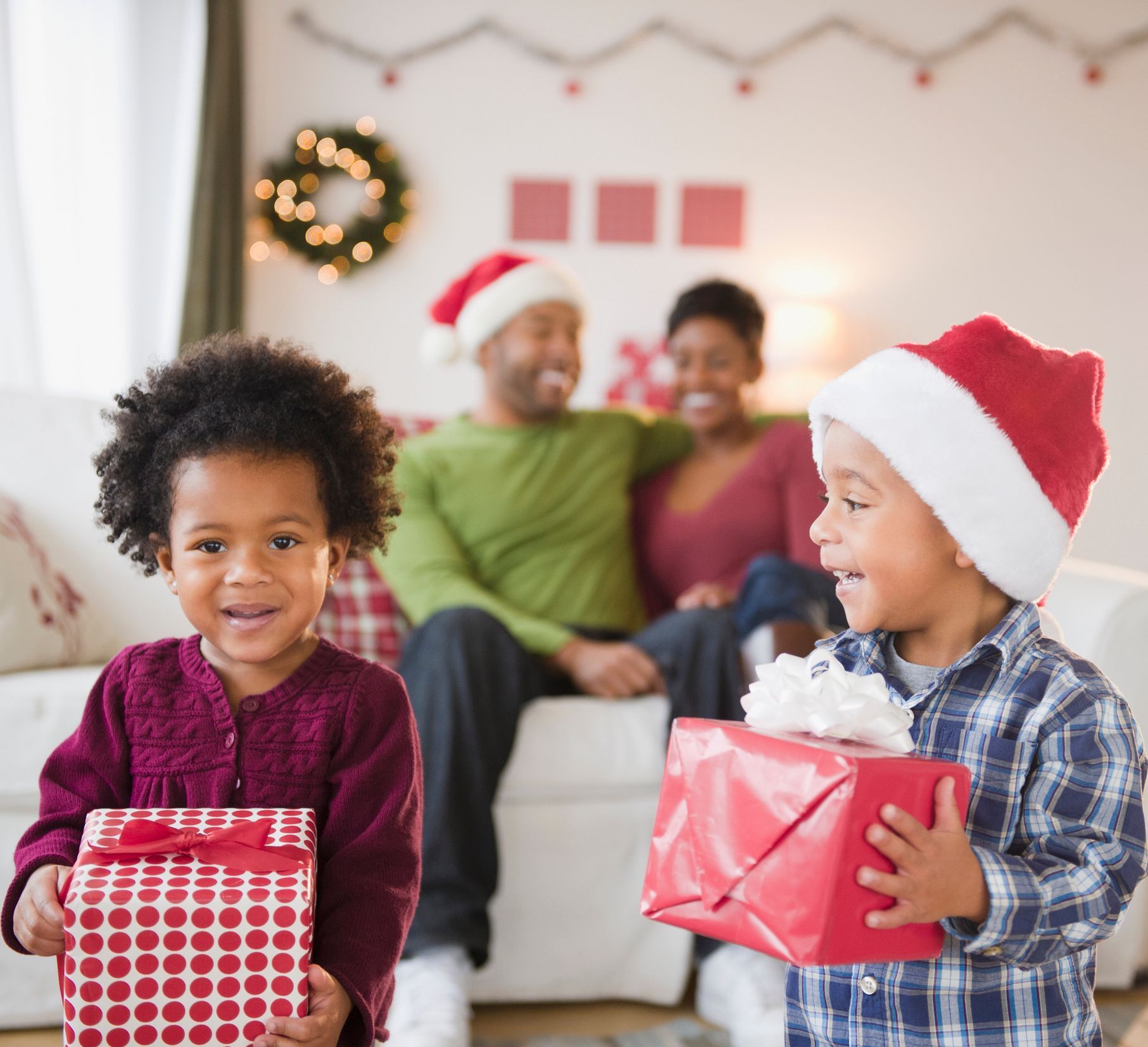 Two children holding Christmas gifts smile in front of their parents on a couch. Two children holding Christmas gifts smile in front of their parents on a couch.