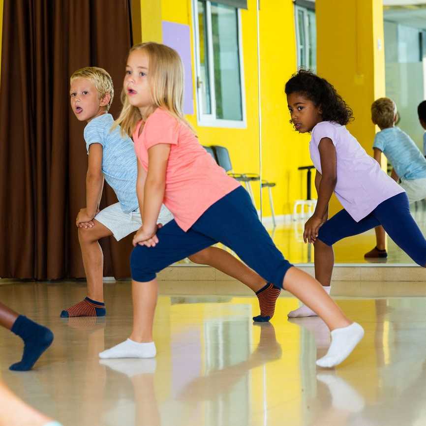Children doing stretching exercises in a dance studio with yellow walls and a mirror.