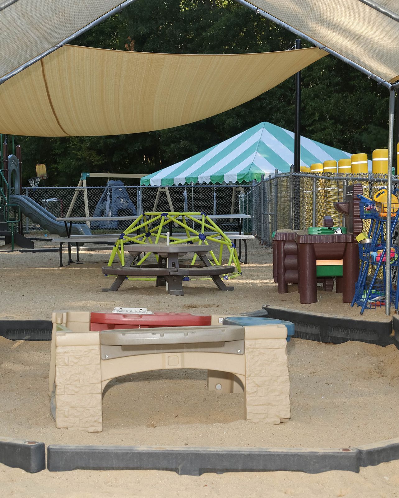 Playground sandbox with shade cover, picnic table, play structures, and tent in the background.