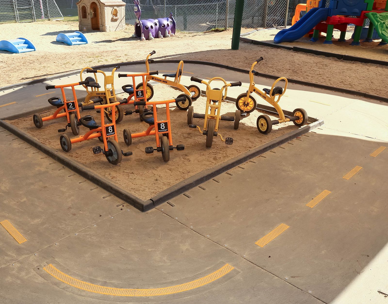 Childcare playground with tricycles on tan ground within a square border. Yellow road markings on concrete.