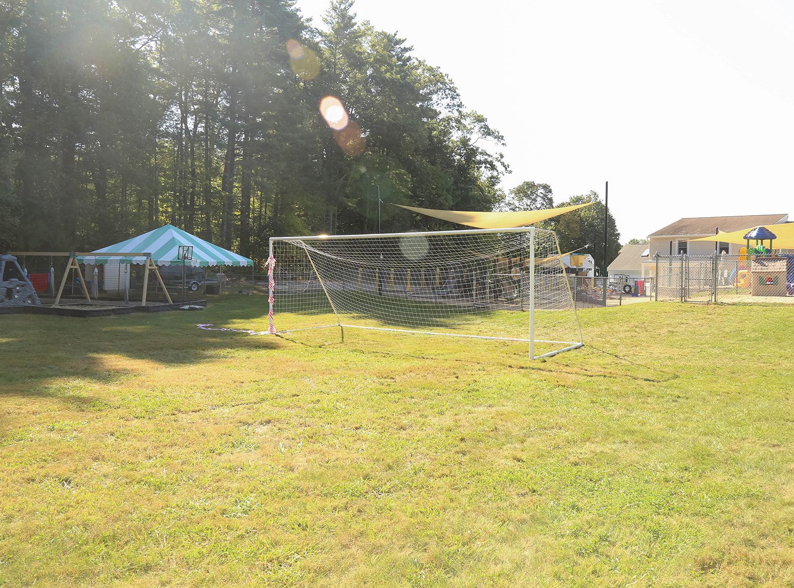 Soccer goal on grassy field, tent, play structure, and trees in the background. Sunny day.