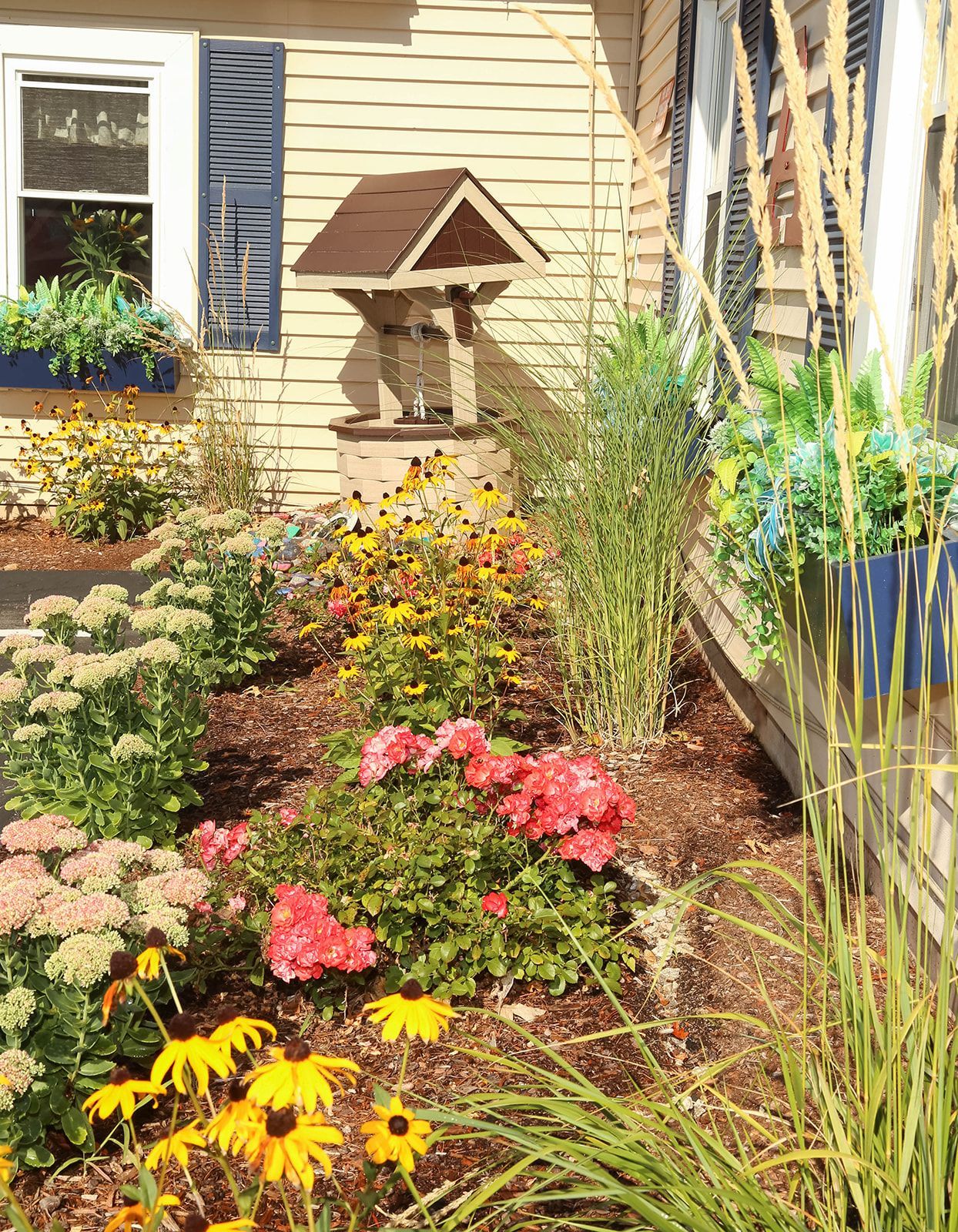 A colorful garden bed with various flowers, a decorative well, and a house with blue shutters.