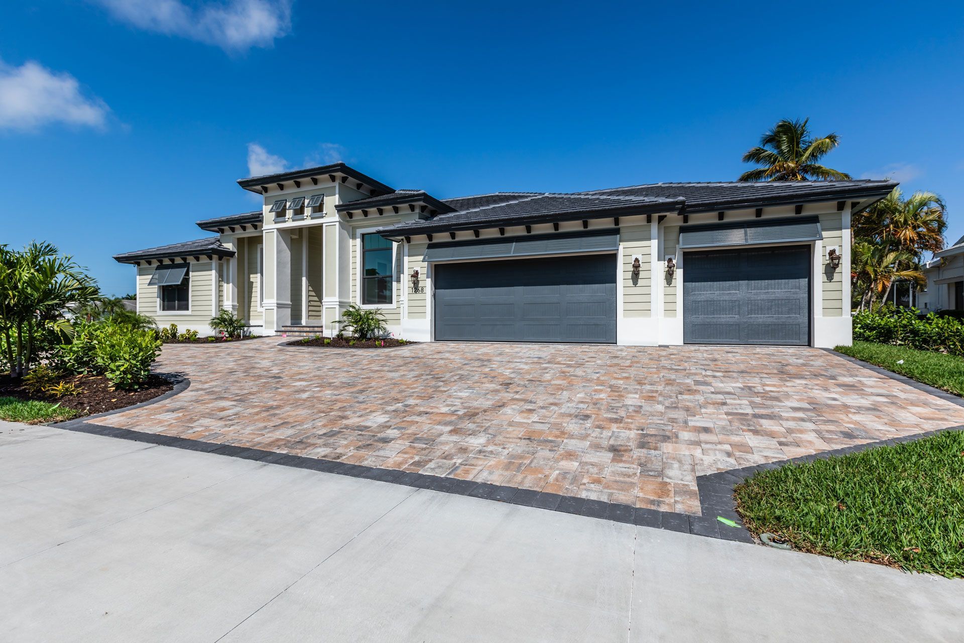 Beige and white luxury home with grey garage doors, brick driveway, and blue sky.