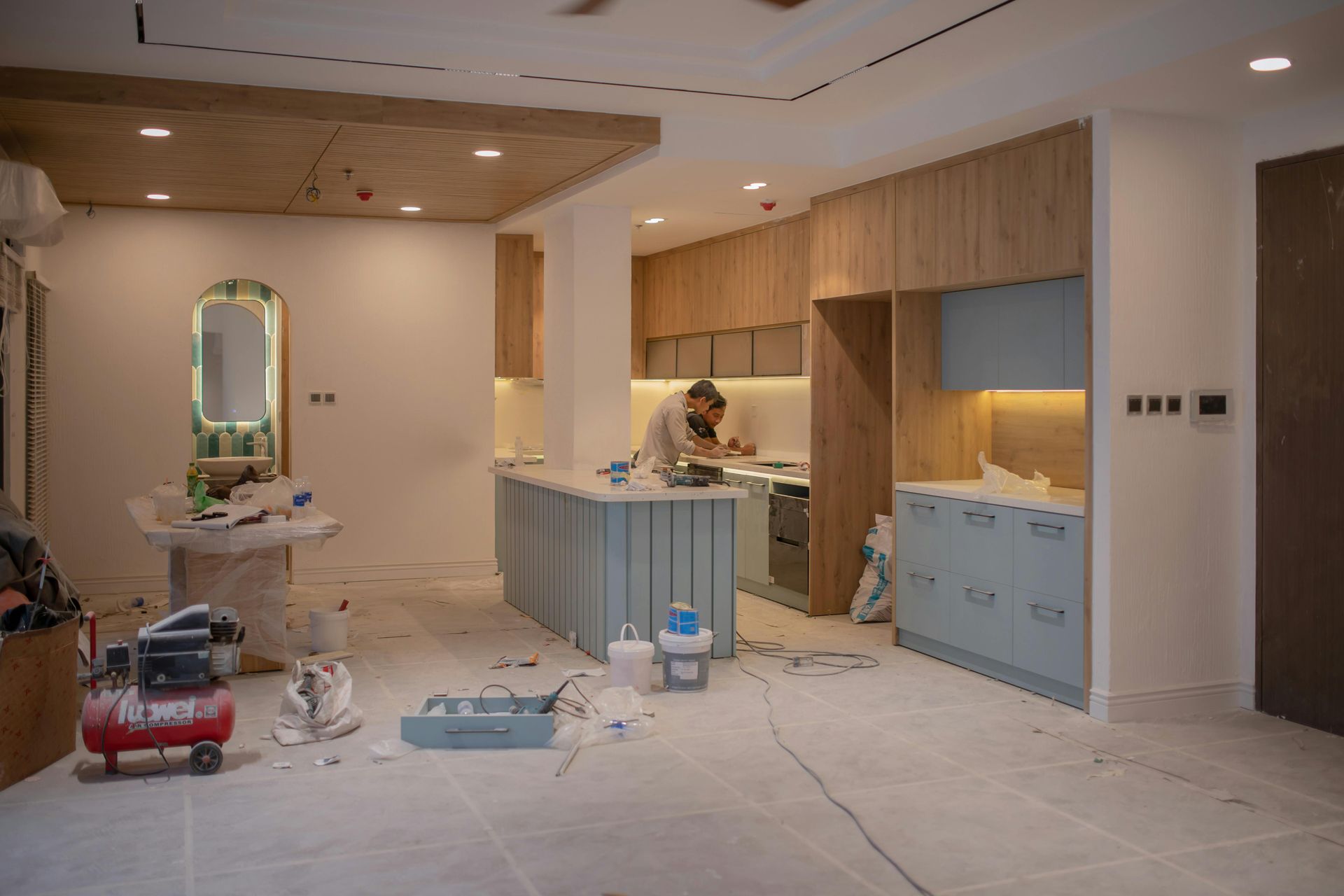 Modern kitchen under construction with workers, unfinished cabinets, tools, and a ladder in a bright room
