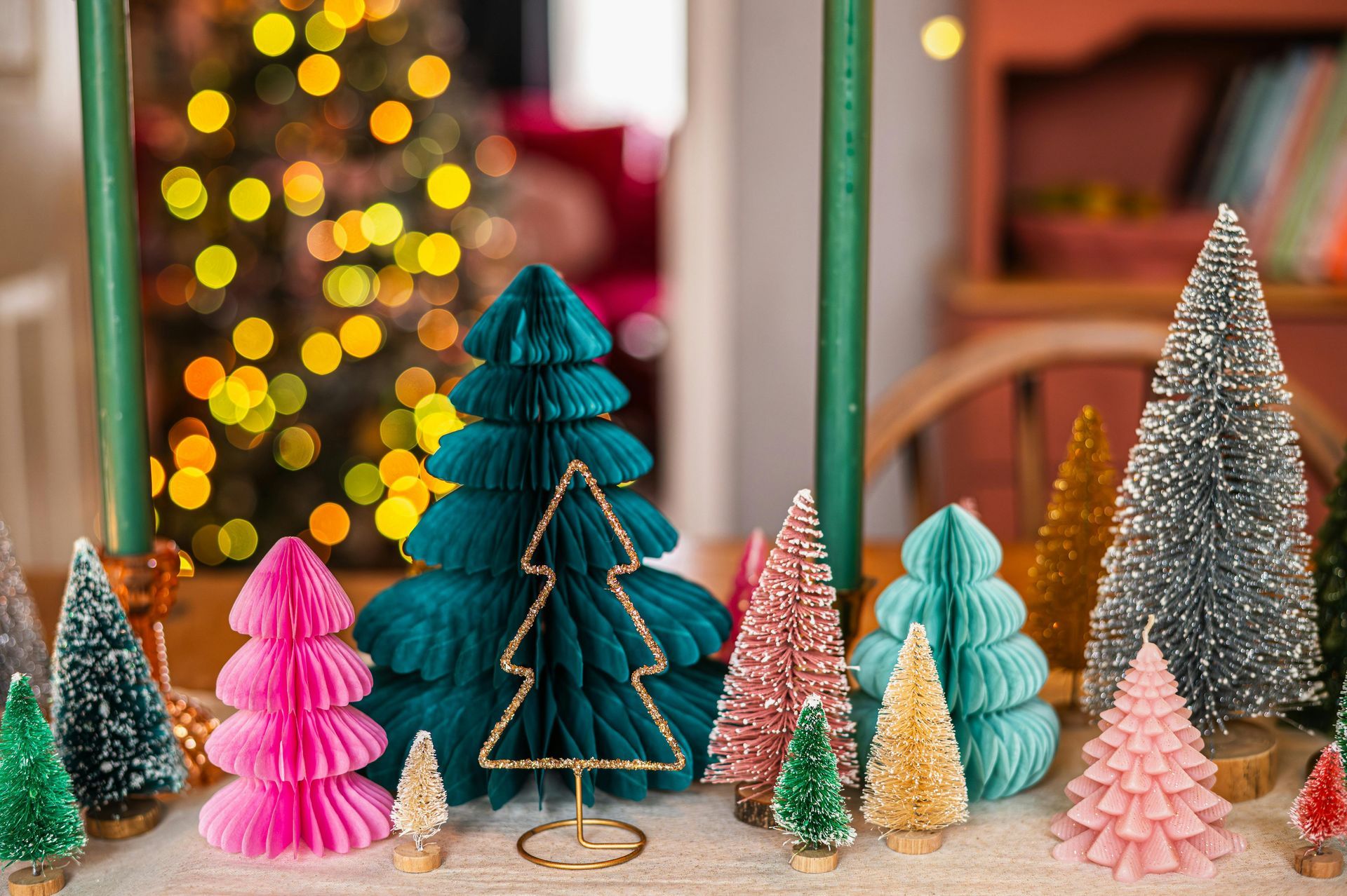 Collection of colorful miniature Christmas trees on a table, with a blurred Christmas tree in the background.