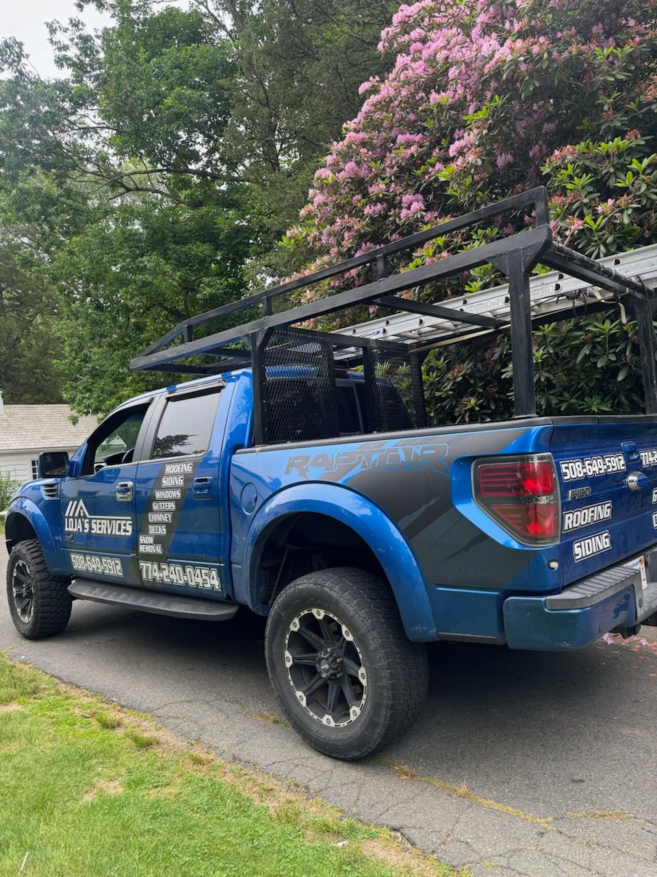 A blue truck is parked on the side of the road next to a bush with pink flowers.