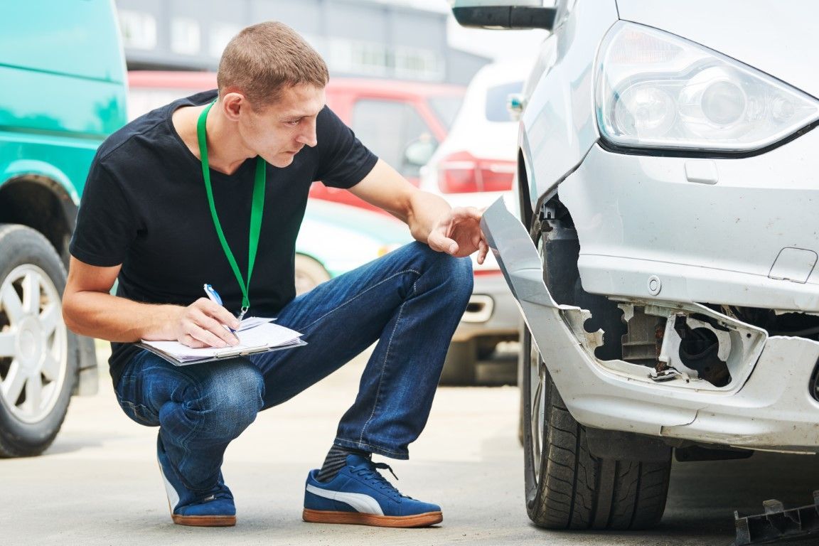 Mechanic crouching beside a silver car, inspecting the front bumper with a clipboard