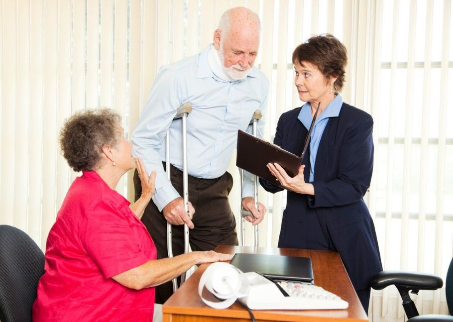 Three people in a meeting, with one woman holding a folder beside an elderly man and seated woman at a table.