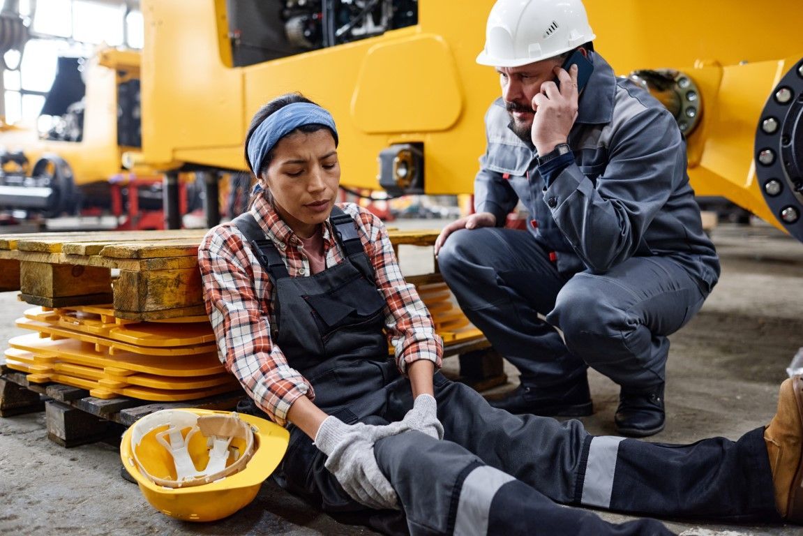 Two workers in hard hats attend an injured colleague on the factory floor with a yellow machine nearby