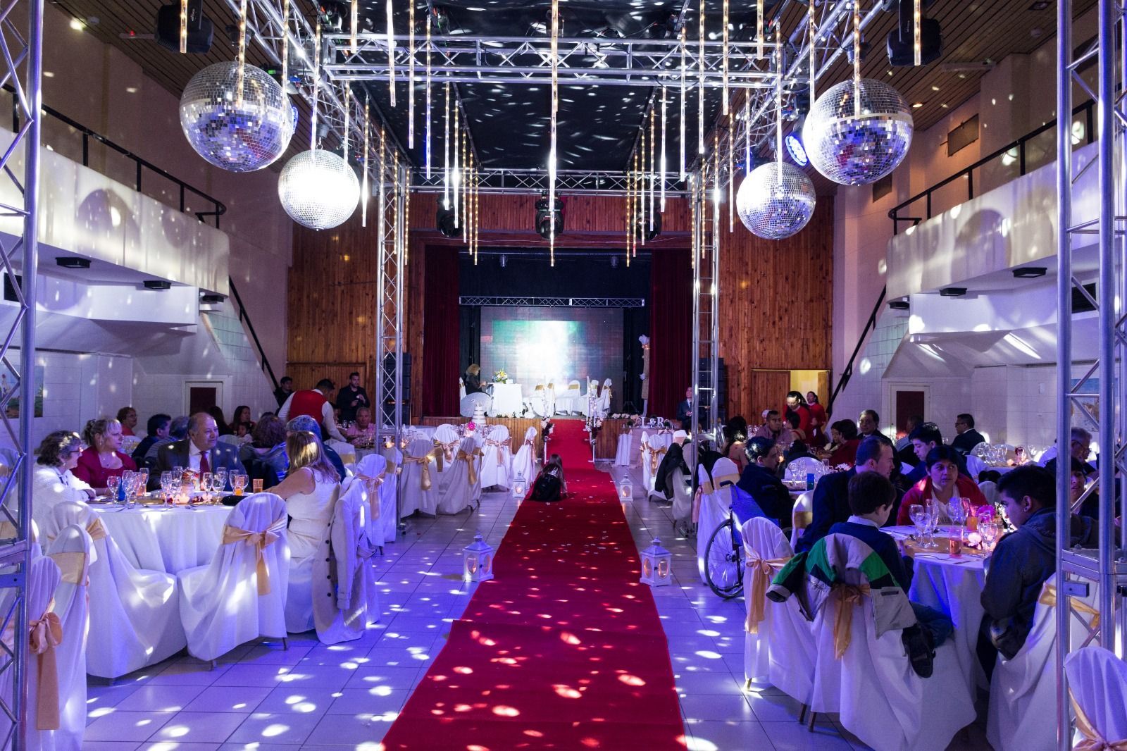 Salón de recepción de bodas con alfombra roja, bolas de discoteca e invitados sentados en mesas.