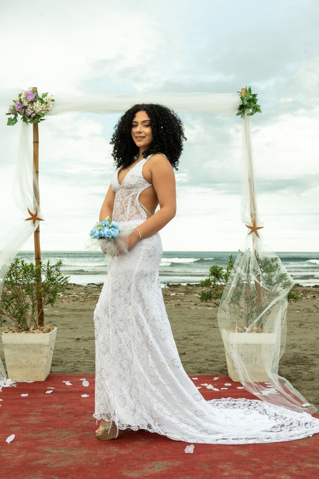 Novia con vestido de novia de encaje blanco sosteniendo un ramo, de pie sobre una alfombra roja en una ceremonia en la playa.