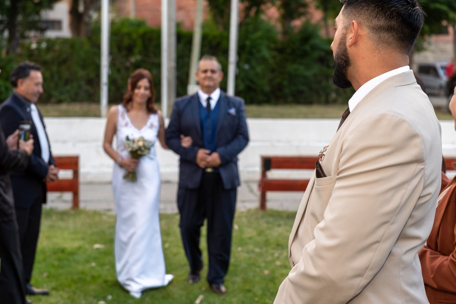 La novia camina hacia el altar con su padre; el novio observa. Ceremonia al aire libre, césped verde, tonos neutros.