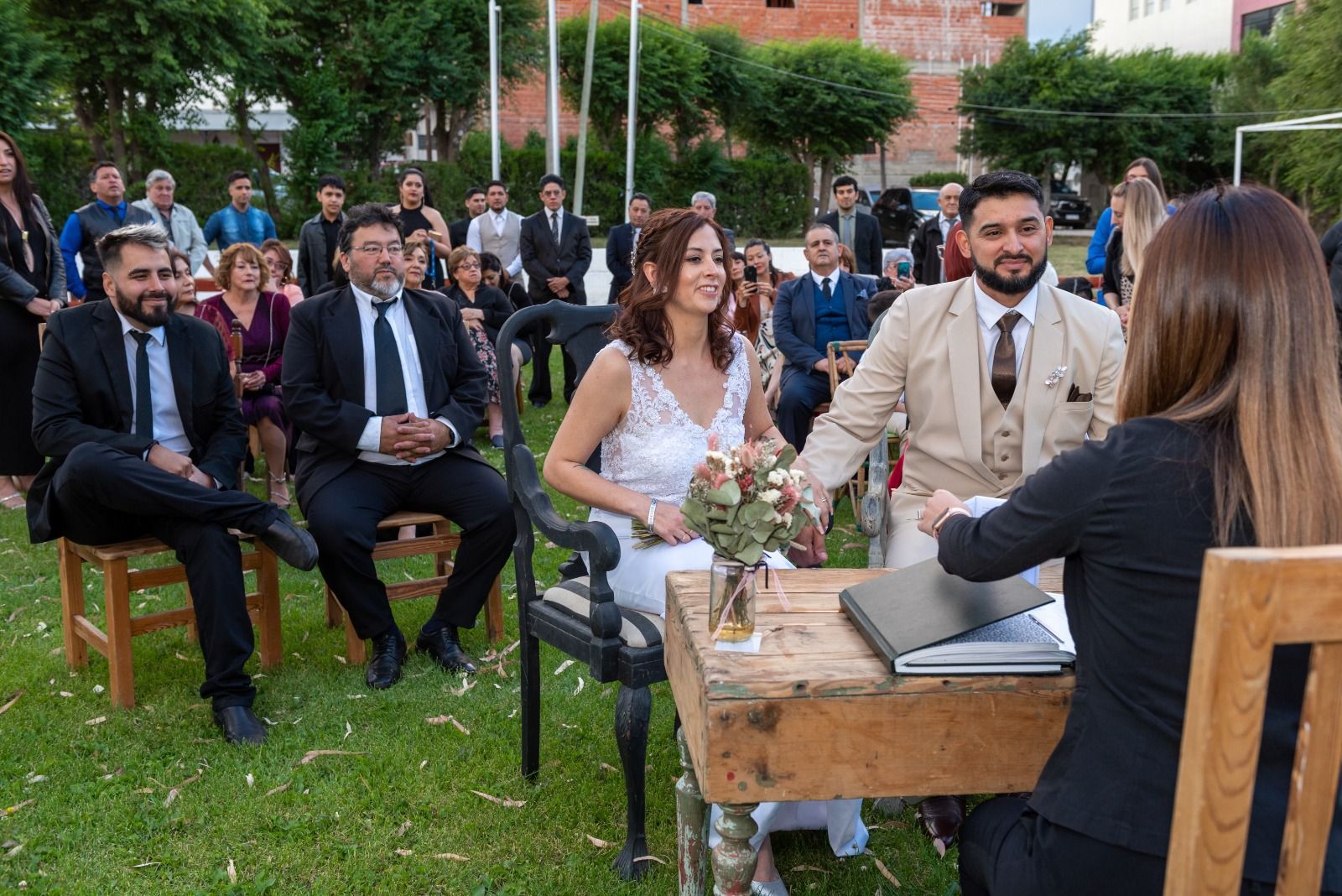 Ceremonia de boda con una pareja firmando documentos en una mesa al aire libre; invitados al fondo.