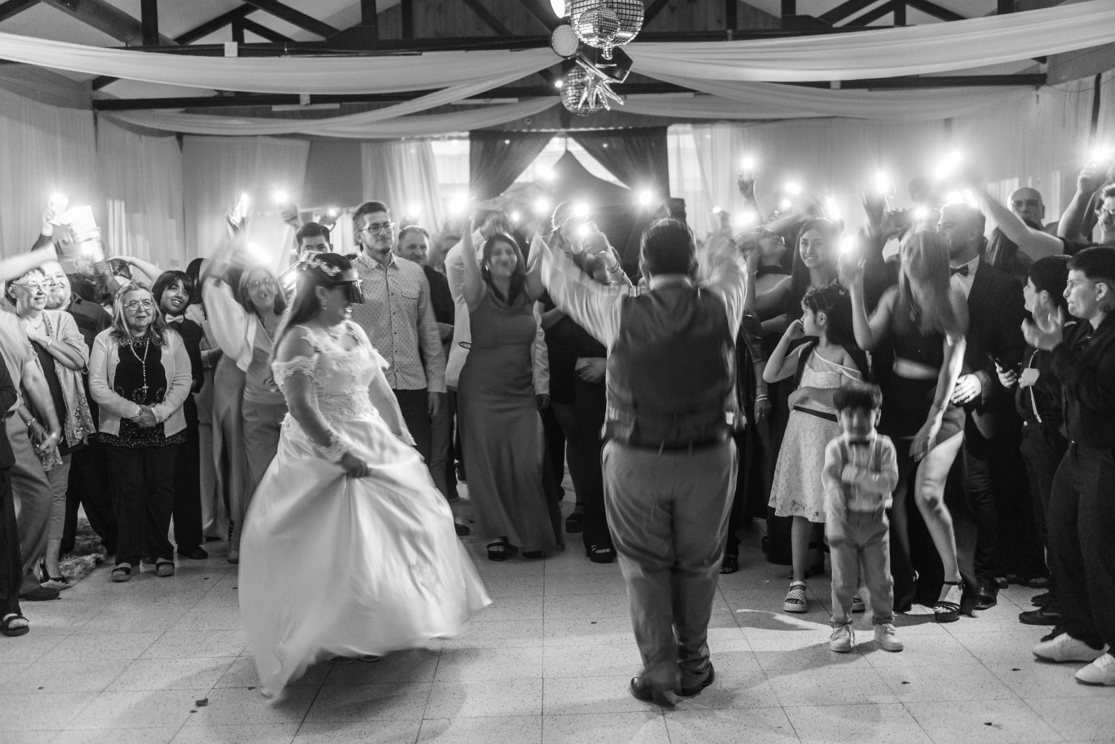 Recepción de boda: novios bailando, invitados sosteniendo teléfonos iluminados, lugar interior.