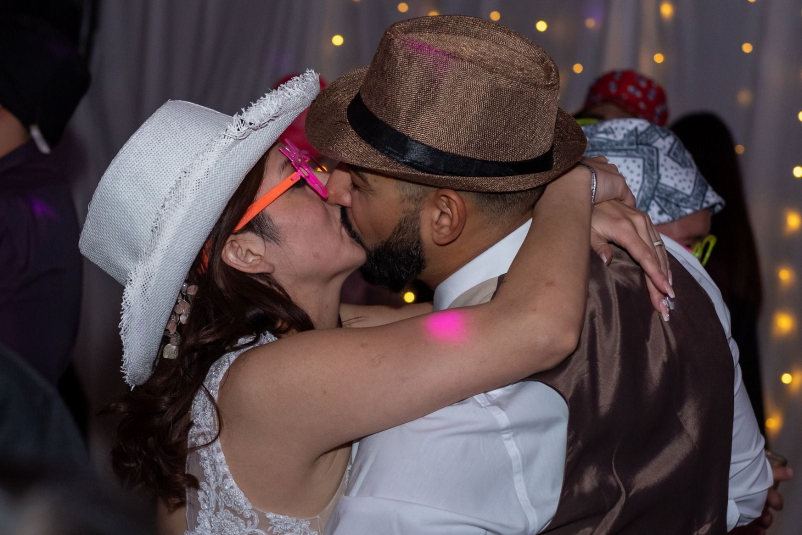 Pareja besándose, con sombreros y gafas de fiesta, con luces de cadena de fondo.