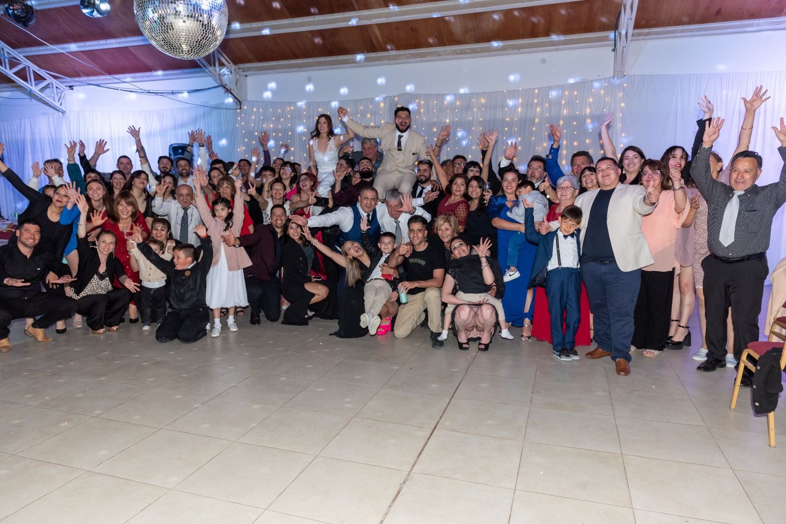Un gran grupo de personas celebrando un evento, muchos de ellos con los brazos en alto. Los novios están en el centro.