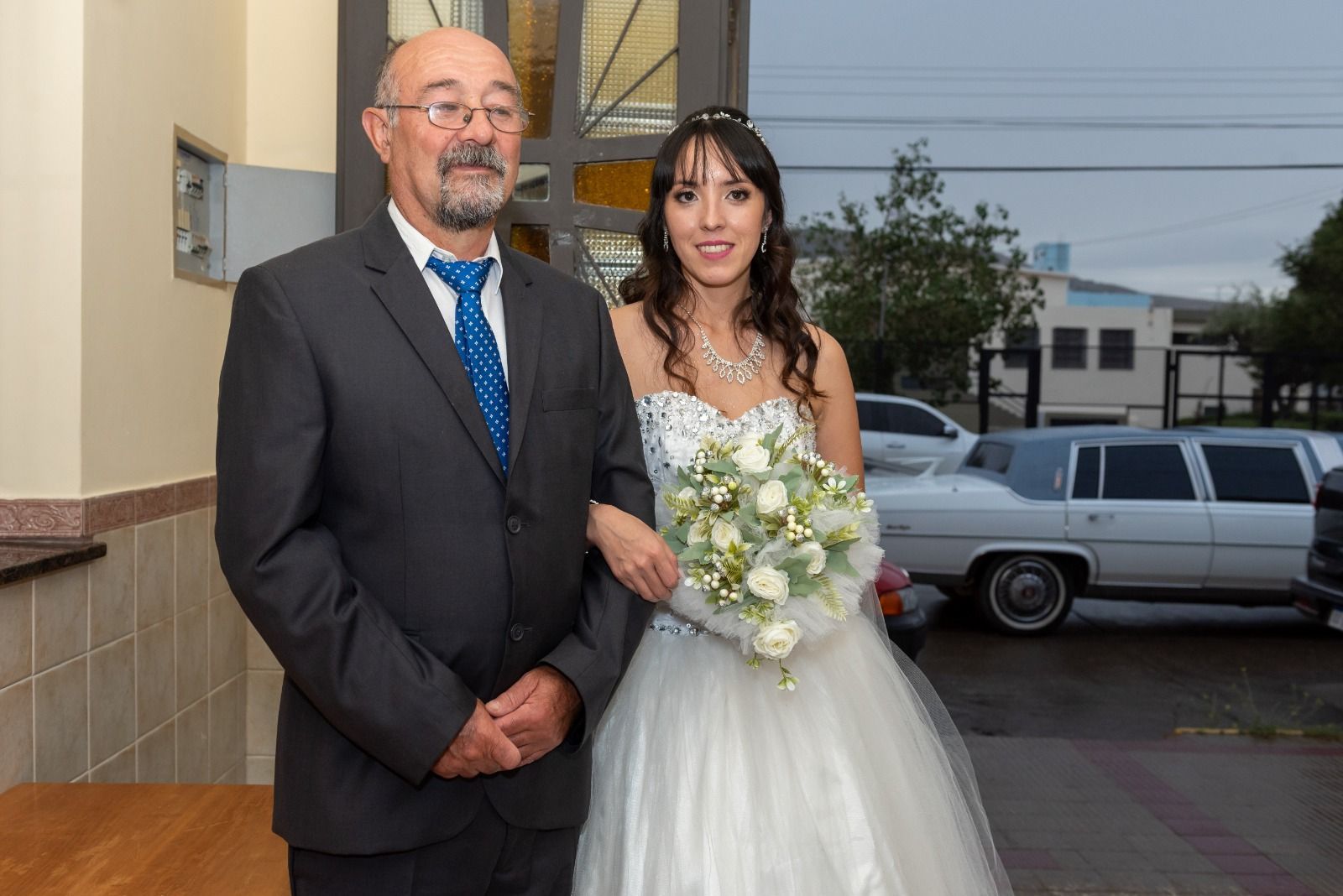 La novia y el hombre de traje salen del edificio; la mujer con vestido blanco lleva un ramo.