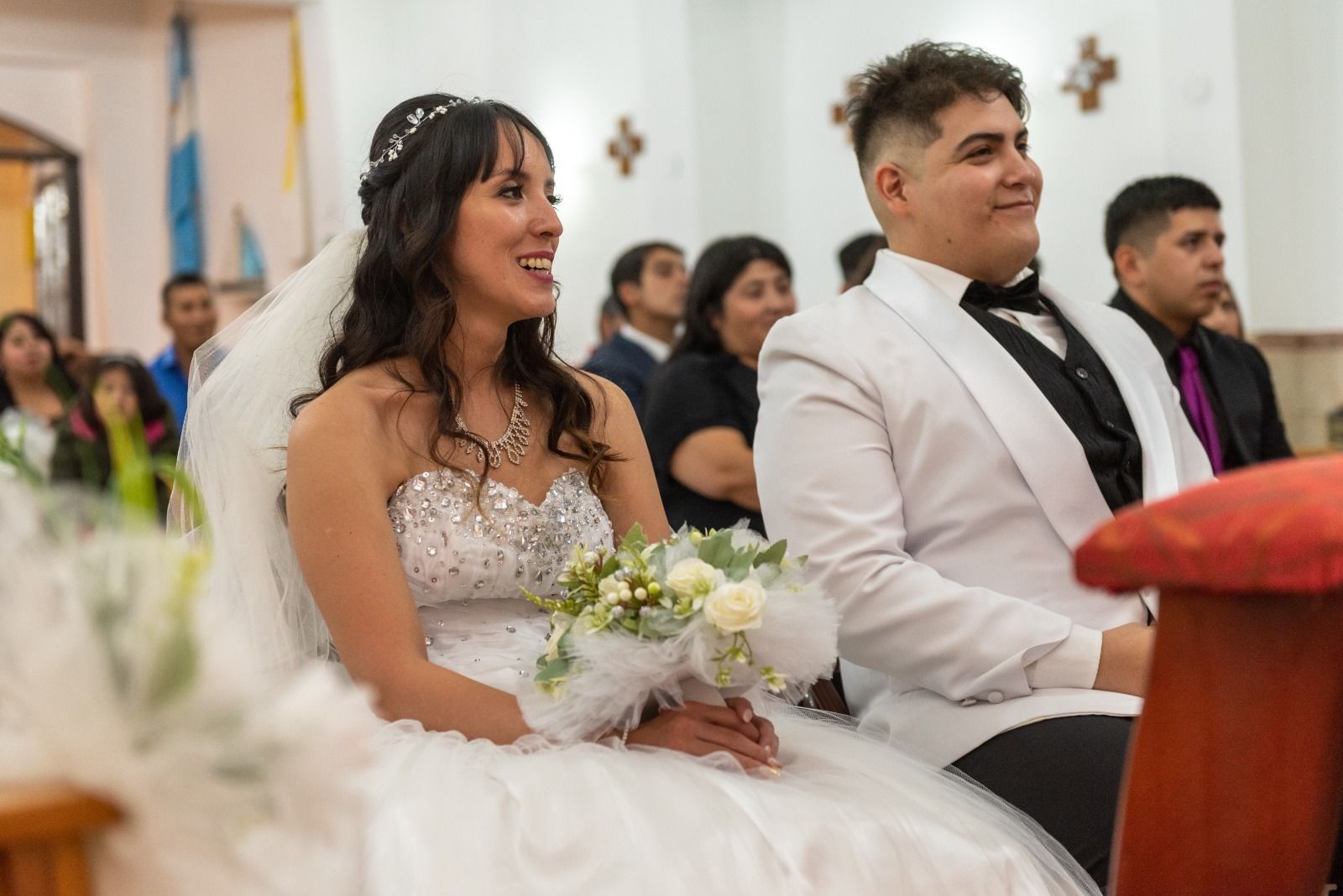 Novios sentados durante una ceremonia nupcial en una iglesia. La novia sonríe, sosteniendo el ramo, y el novio observa.