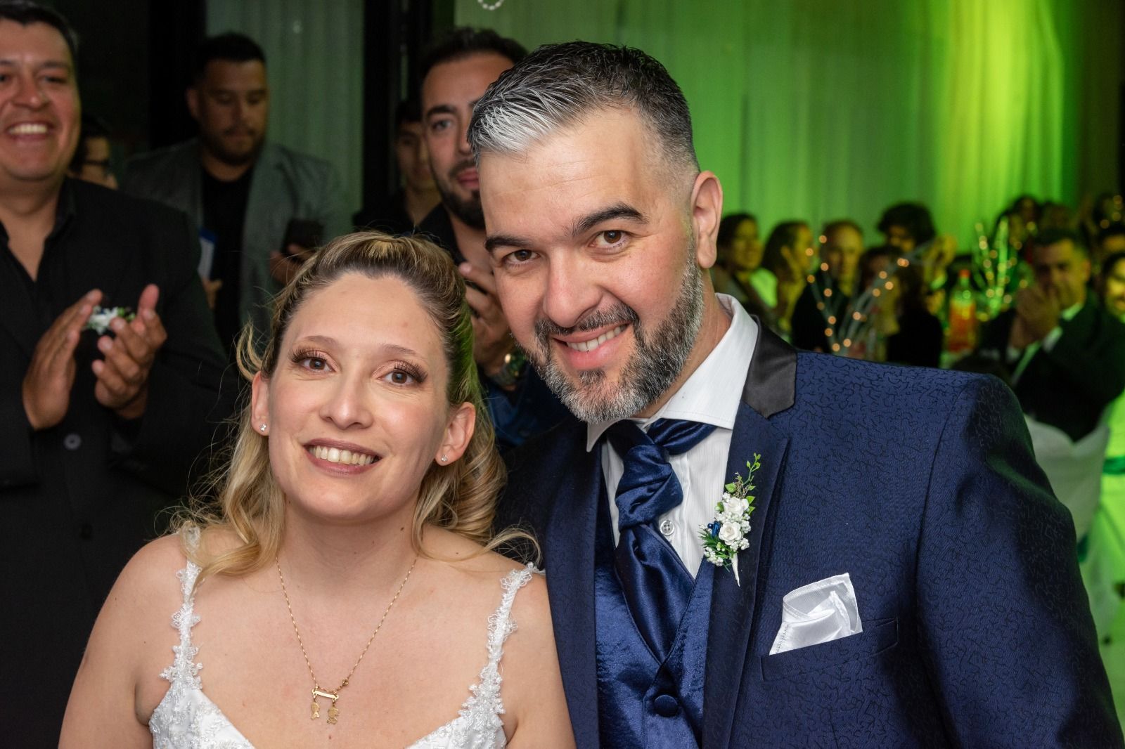 Novia y novio sonriendo en una boda, él con un traje azul, ella con un vestido blanco, otros al fondo.