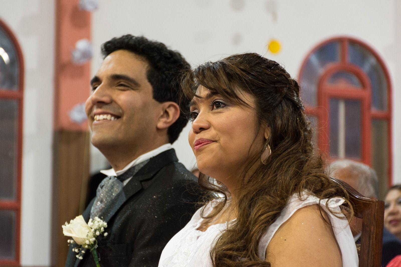 Pareja sonriente en una ceremonia de boda; el hombre lleva un traje con un boutonniere y la mujer lleva un vestido blanco.