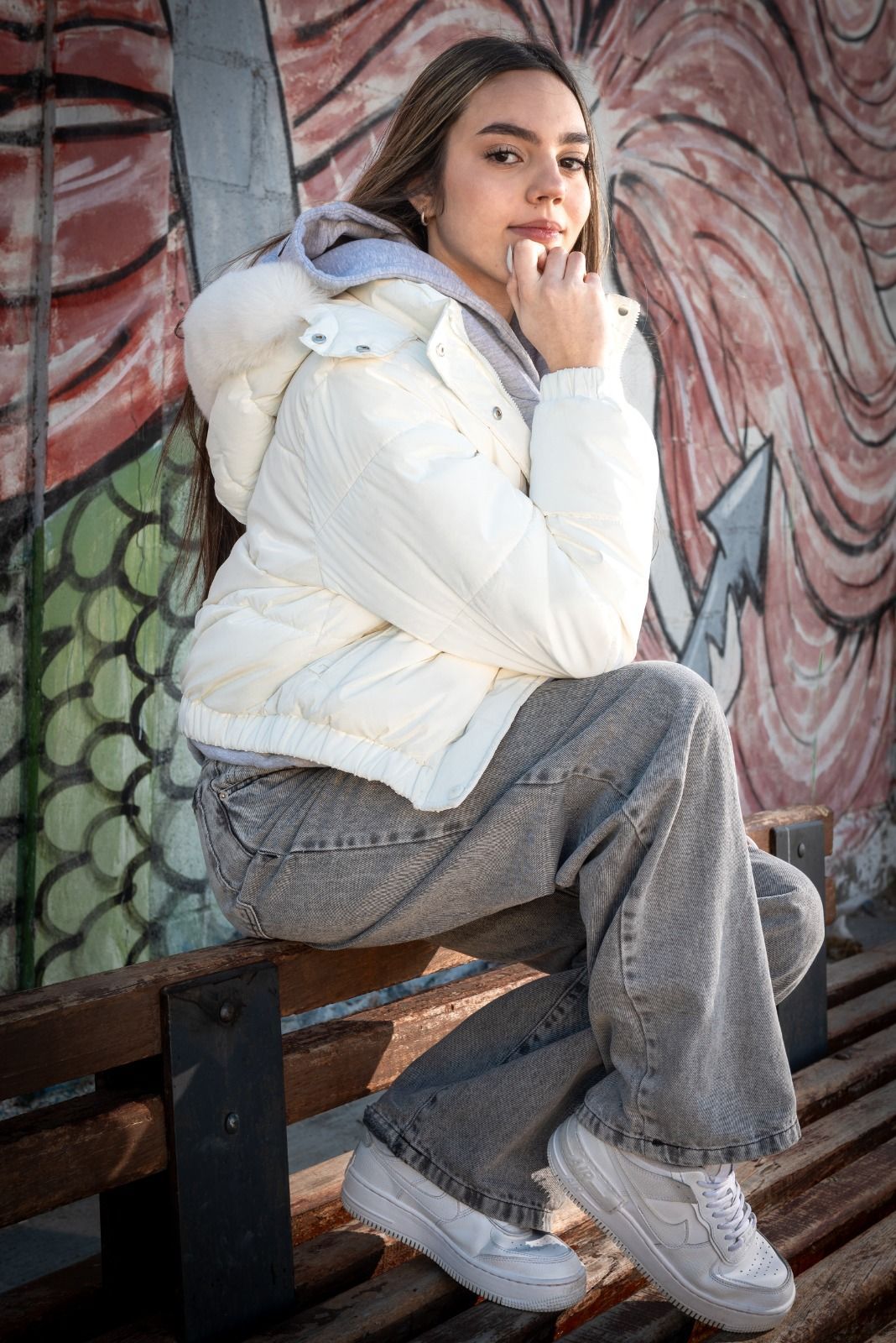Una mujer con una chaqueta acolchada blanca y jeans grises se sienta en un banco con un grafiti de fondo.